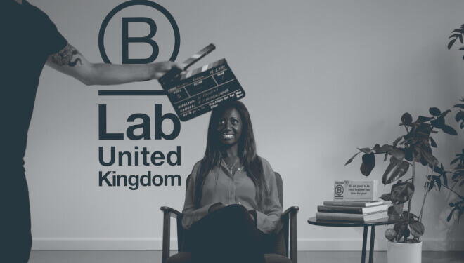 A woman seated and smiling as a clapperboard is held above her, with the 'B Lab United Kingdom' logo, a table with books, a plant, and a certification sign in the background.