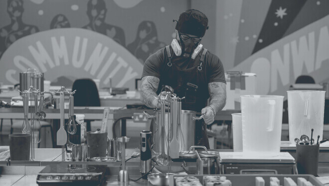 A person wearing an apron and protective gear works with brewing equipment at a workstation, set against a colourful mural featuring the word 'Community' in the background.