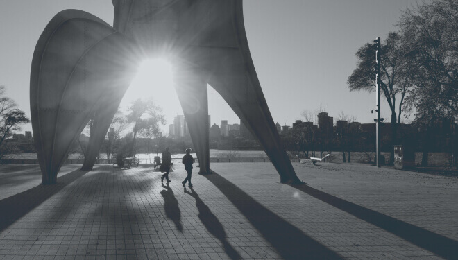 Silhouettes of people walking under a large sculpture at sunset, with long shadows stretching across the tiled ground and a city skyline in the background.