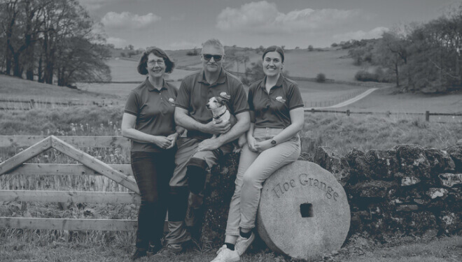 Three people wearing matching purple polo shirts stand by a stone wall engraved with 'Hoe Grange,' posing with a small dog, set against a backdrop of green fields and a cloudy sky.