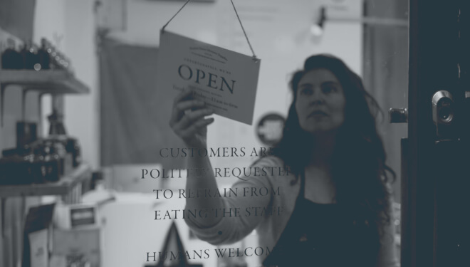 A woman hanging an 'Open' sign on a glass door with humorous text: 'Customers are politely requested to refrain from eating the staff. Humans welcome.'