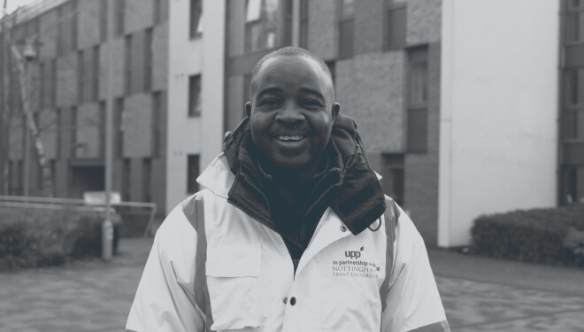 A smiling man wearing a high-vis jacket with a logo stands outdoors in front of modern brick and concrete buildings.